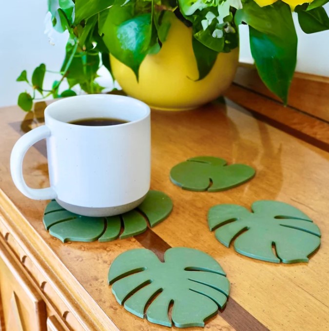 A white mug filled with coffee sits on a wooden surface, accompanied by three green leaf-shaped coasters. In the background, there is a yellow pot with green leaves, creating a cozy and inviting atmosphere.