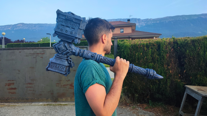 A man in a green shirt is holding a large, intricately designed hammer-like object on his shoulder, with a mountainous landscape in the background.