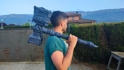 A man in a green shirt is holding a large, intricately designed hammer-like object on his shoulder, with a mountainous landscape in the background.