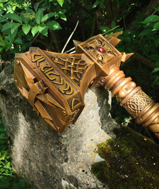 A golden, ornate sword with intricate designs and a red gemstone on top, resting on a mossy rock in a lush, green environment.