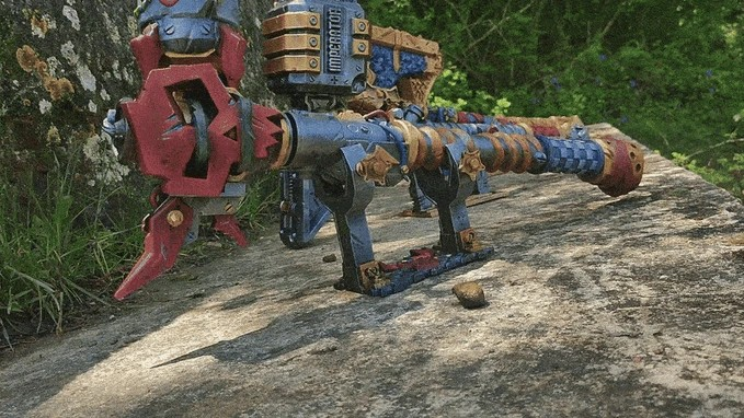 A colorful toy robot with a red and blue body and various mechanical components, including a large head and a long body, is situated on a concrete surface surrounded by greenery.