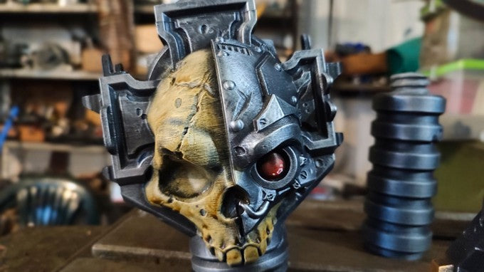 A close-up view of a skull-shaped object with a metallic, futuristic design, featuring a red eye and a metallic, angular face.