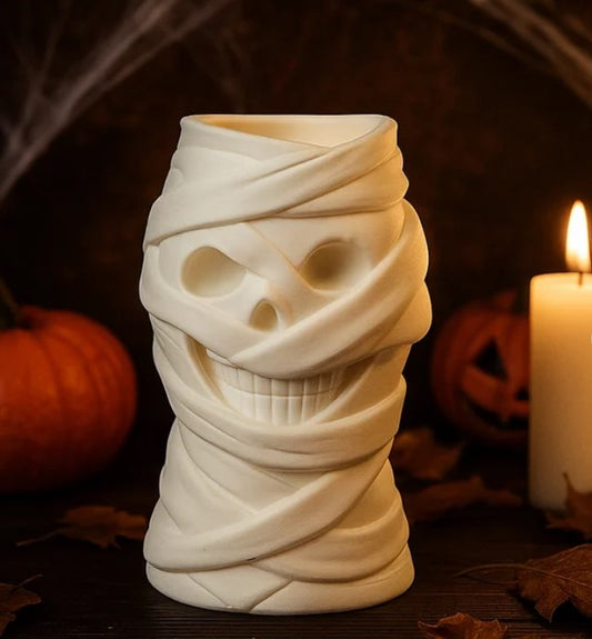 A white ceramic mug with a mummified skull design, resembling a mummy wrapped in bandages, is placed on a table with a lit candle and pumpkins in the background.