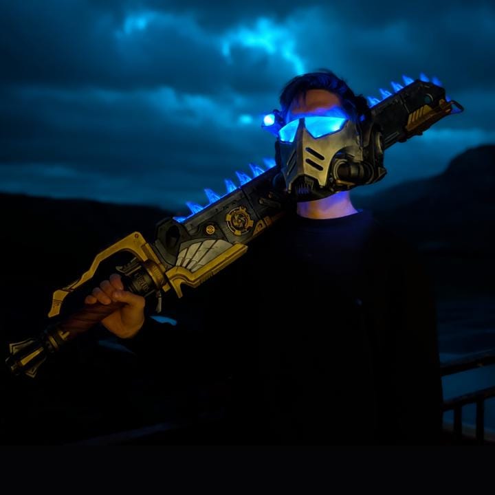 A person wearing a futuristic-looking mask and holding a large, glowing sword-like weapon stands in a dark, cloudy environment.