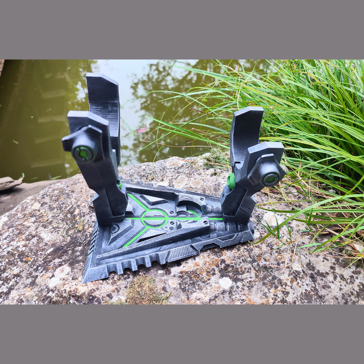 A 3D printed object resembling a futuristic device or tool is placed on a rock near a body of water, surrounded by green grass.