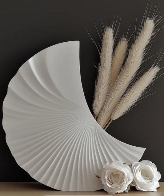 a white vase with a fan-like design, accompanied by a bouquet of white flowers and a few long, thin stems.
