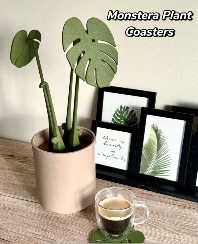 A potted monstera plant with green leaf-shaped coasters sits on a wooden surface, accompanied by a cup of coffee and framed plant prints.