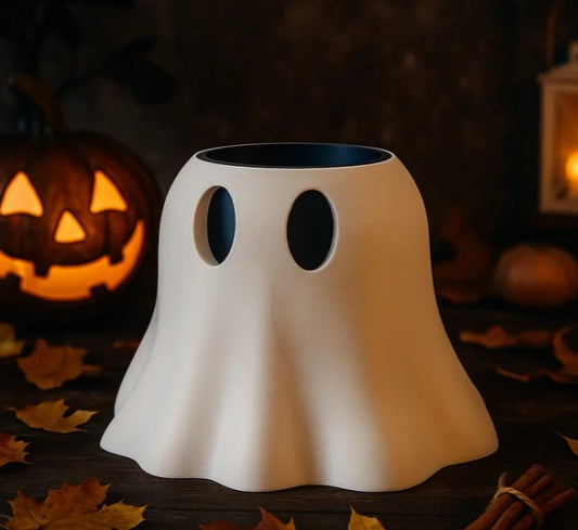 A white ghost-shaped container with two holes on the front sits on a wooden surface, with a carved jack-o-lantern and autumn leaves in the background.