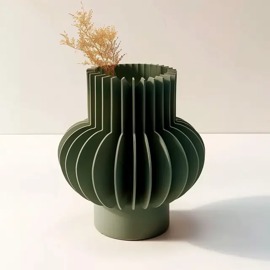 A modern, sculptural green vase with a unique, ribbed design sits on a white surface, featuring a small plant in the top left corner.