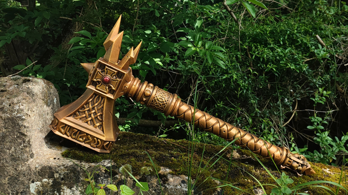 A golden, ornate sword or weapon with a pointed blade and decorative patterns rests on a mossy rock in a lush, green forest setting.