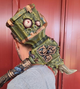 A person wearing a green and brown mechanical device with a skull-like design on the back, standing in front of a red wall.