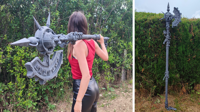 A woman in a red top and black pants is holding a large, futuristic-looking weapon in a wooded area.