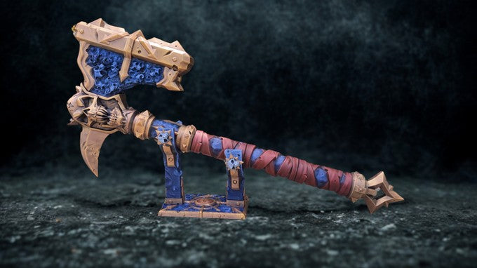 A highly detailed and ornate fantasy weapon, possibly a sword or axe, with intricate designs and patterns in blue and gold colors. The weapon appears to be a collectible item or a decorative piece, displayed on a stand against a dark background.