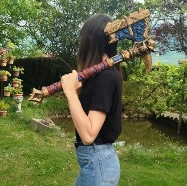 A woman is standing in a garden, holding a large, ornate sword-like weapon with a blue and gold design.