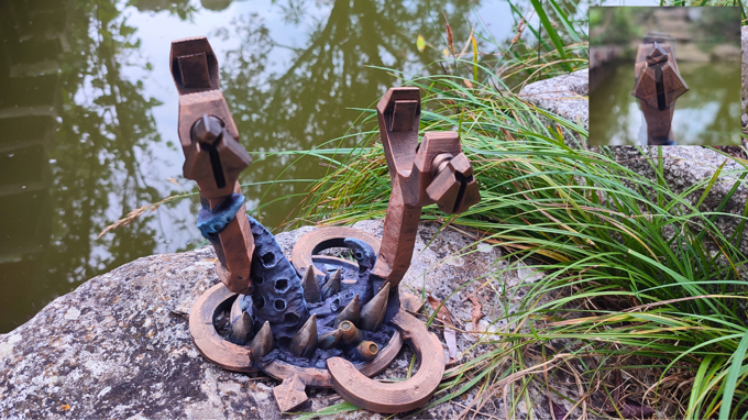 a unique, abstract sculpture composed of various shapes and textures, including a combination of wood and metal elements, situated on a rock near a body of water.