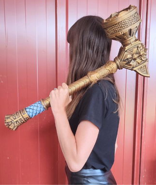 A woman with long brown hair is holding a large golden object, which appears to be a weapon or tool, in front of a red wall.